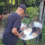 A person wearing a black shirt and cap plays a Panworld steel pan drum in a garden surrounded by greenery and outdoor furniture.