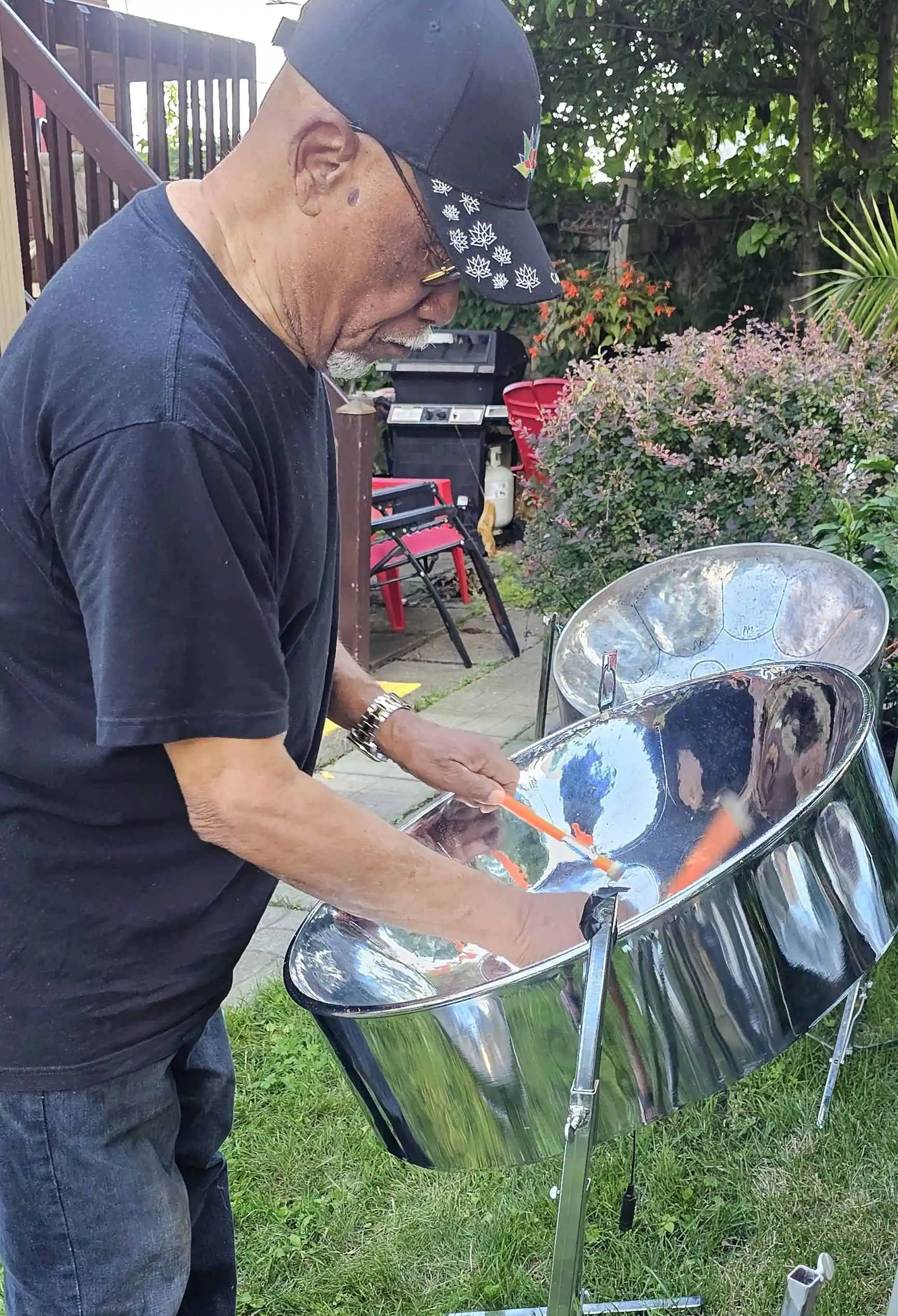 An older man wearing a cap and glasses plays a steel pan drum for sale outdoors in a beautiful garden setting.