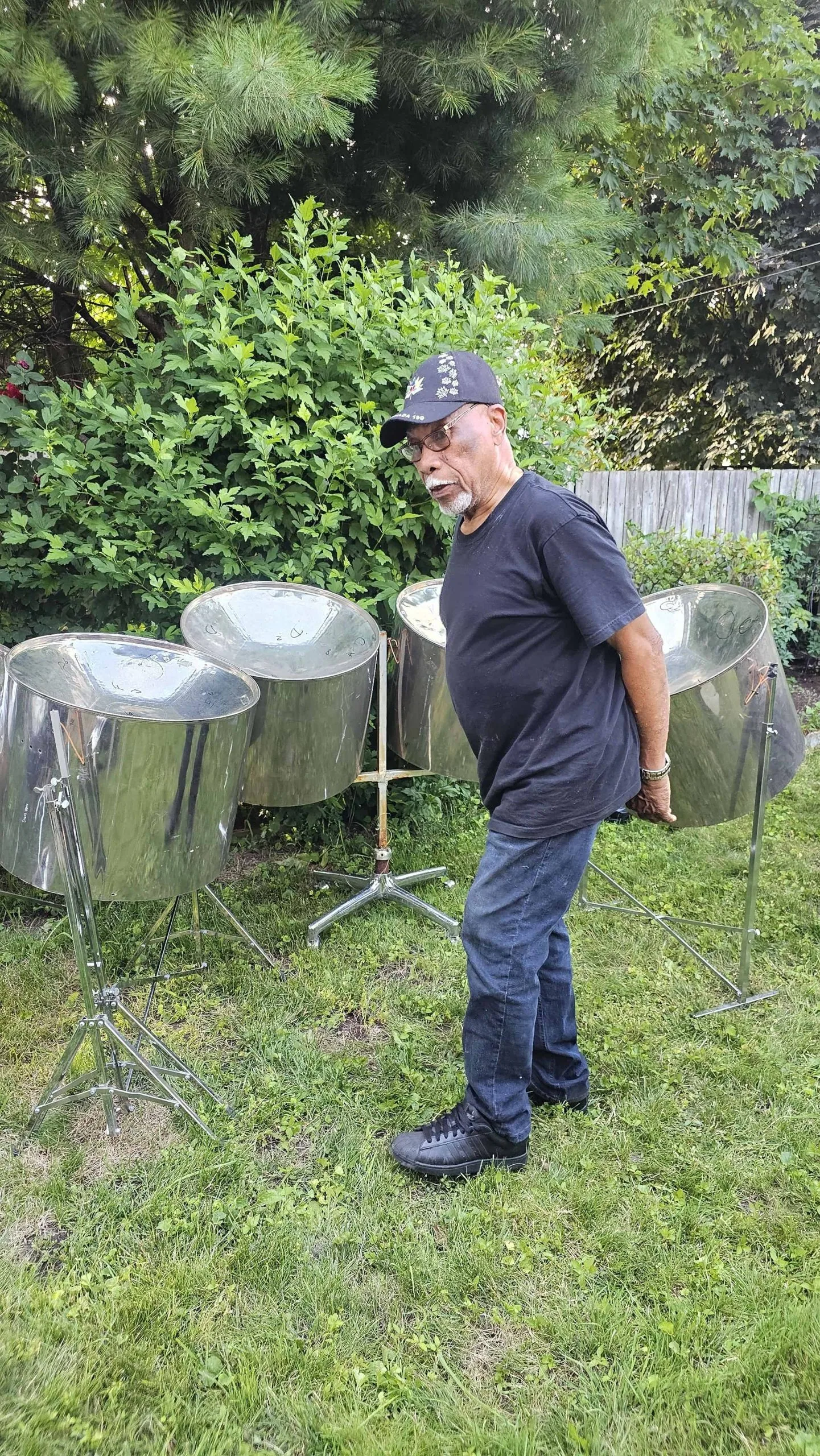 An older man in a black shirt, jeans, and cap stands outdoors on grass beside three steel drums with green bushes in the background, showcasing steel pans for sale.