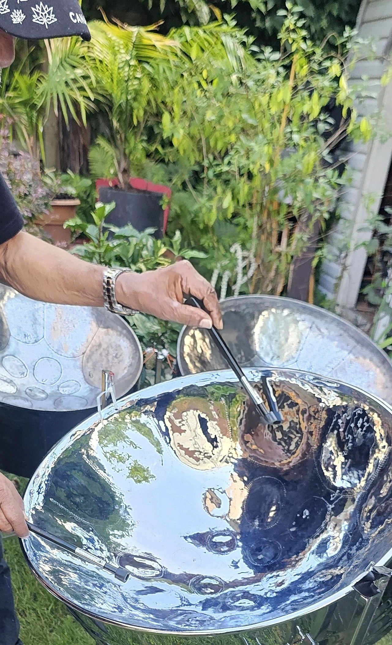 A person playing a shiny steel pan drum for sale outdoors, surrounded by lush green plants and foliage.