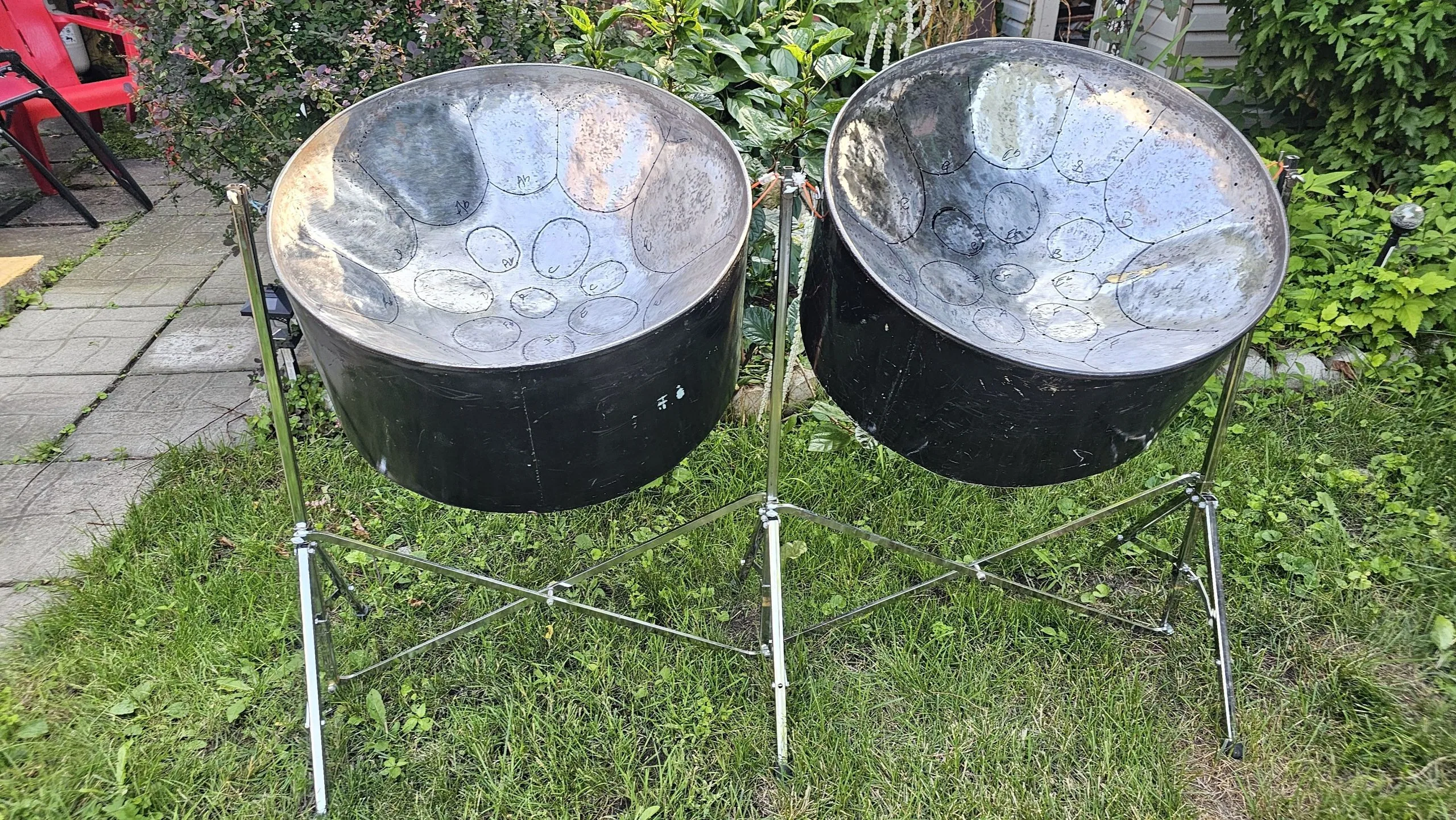 A pair of steelpan drums on a metal stand set on grass, with plants and patio tiles visible in the background—perfect for those seeking quality Steel Pans for Sale.