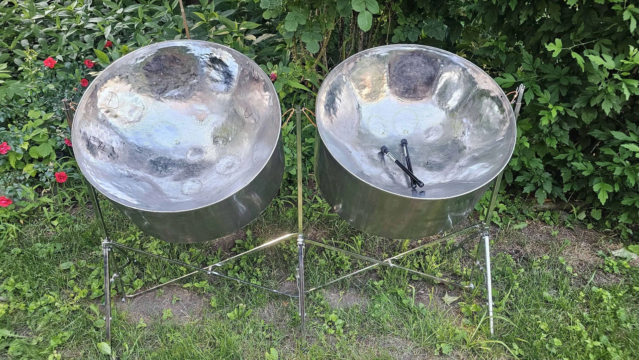 Two steel pan drums on metal stands are set on grass outdoors, surrounded by greenery and flowering plants. Mallets rest on the drum on the right, creating a serene scene—perfect for anyone searching for steel pans for sale.