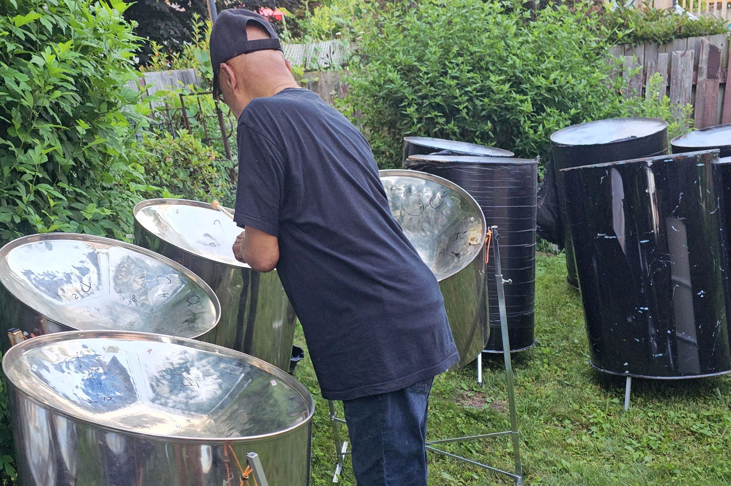 A person wearing a black shirt and cap inspects a set of steel drums in a grassy backyard, showcasing Steel Pan Tuning by Master Tuner James Andrews | PanWorld, surrounded by lush greenery and other drums.